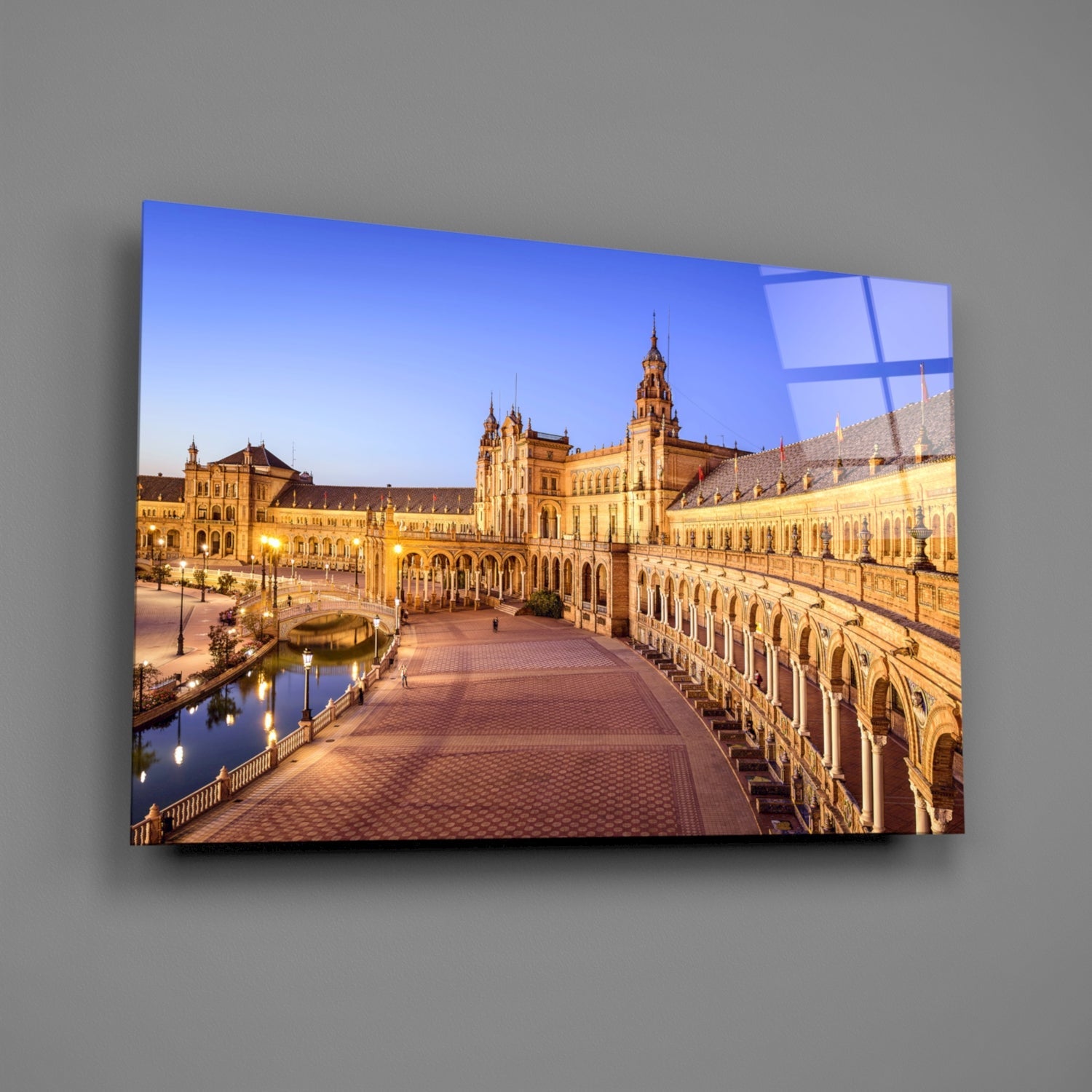 Illuminated Plaza de España in Seville displayed on a glass wall art panel mounted on gray wall, showing golden architecture and blue twilight sky.