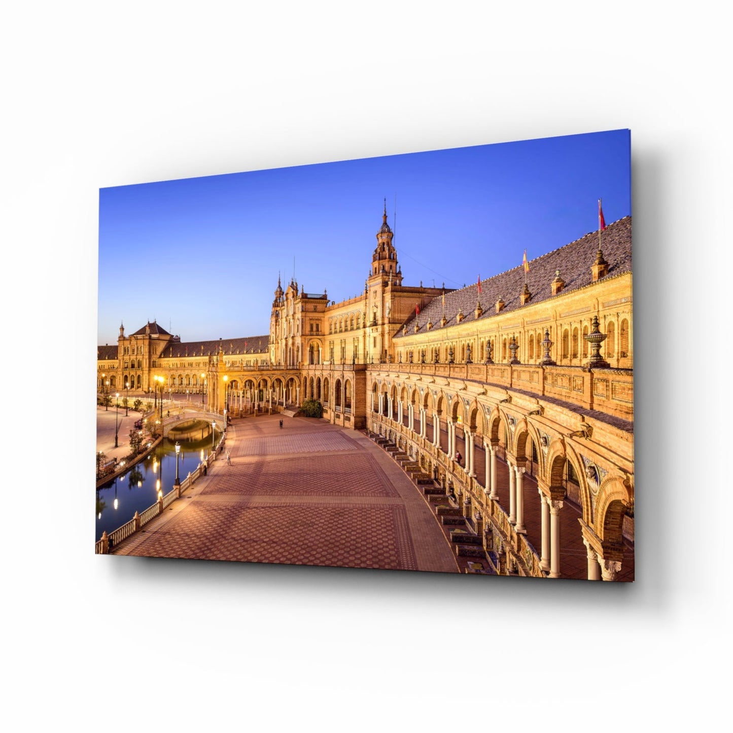 Evening view of Plaza de España in Seville showing illuminated Renaissance Revival architecture with arched colonnade and central tower reflected in canal water.