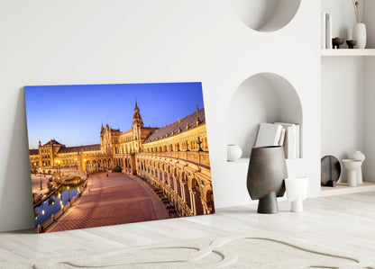 Glass wall art leaning against white wall displays illuminated Spanish plaza architecture with golden buildings and blue evening sky.