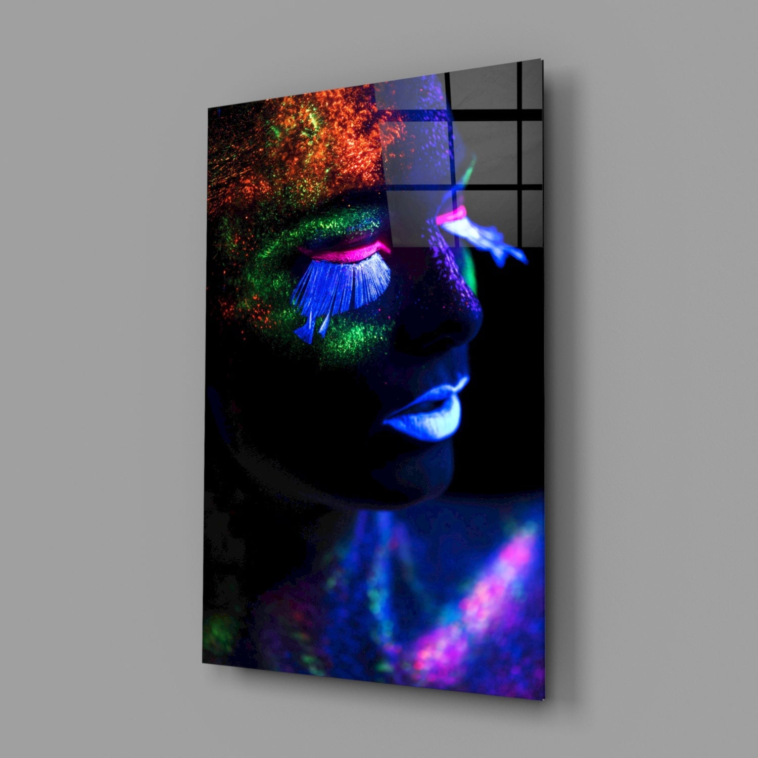 Angled 3D product view of neon makeup glass wall art showing a face with vibrant blue, pink, green, and orange fluorescent paint effects against black background.