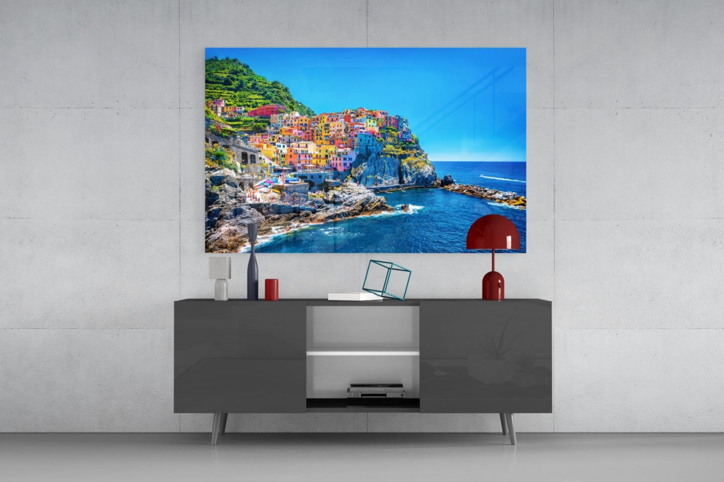 Colorful coastal village glass wall art displayed above dark gray media console with red decorative accessories.