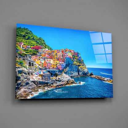 Colorful cliffside village with blue sea reflected on glass wall art mounted on gray wall, angled perspective view.