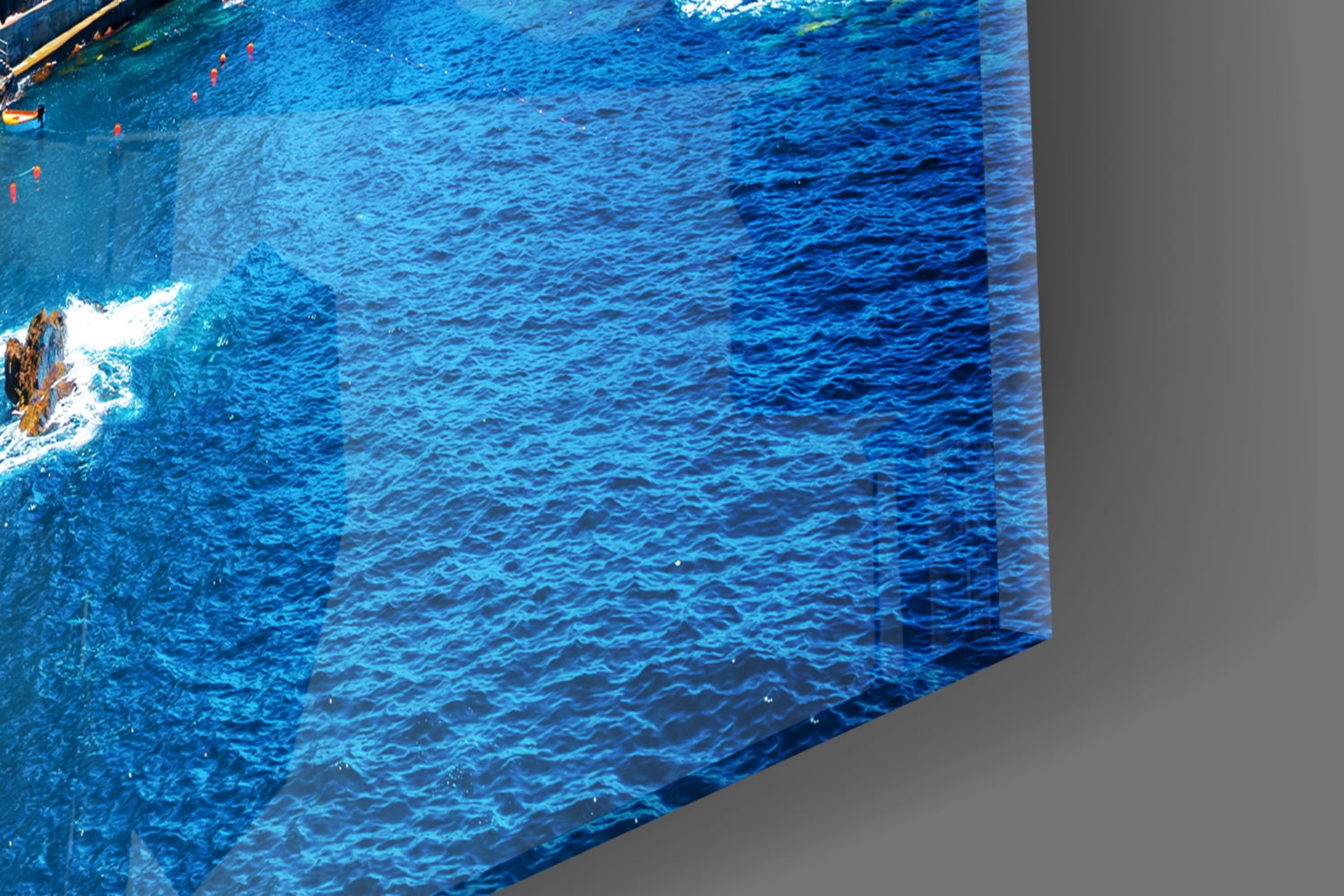 Glass wall art panel displaying aerial view of bright blue ocean water with white foam and rocky coastline along left edge.