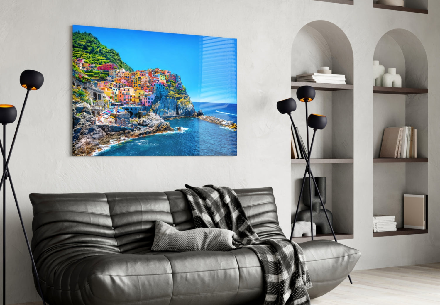 Colorful Italian coastal village with vibrant houses on rocky cliffs overlooking blue sea, displayed as glass wall art above black leather sofa.