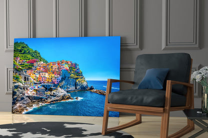 Colorful coastal village glass wall art depicting Manarola, Italy displayed in modern living room next to wooden chair.