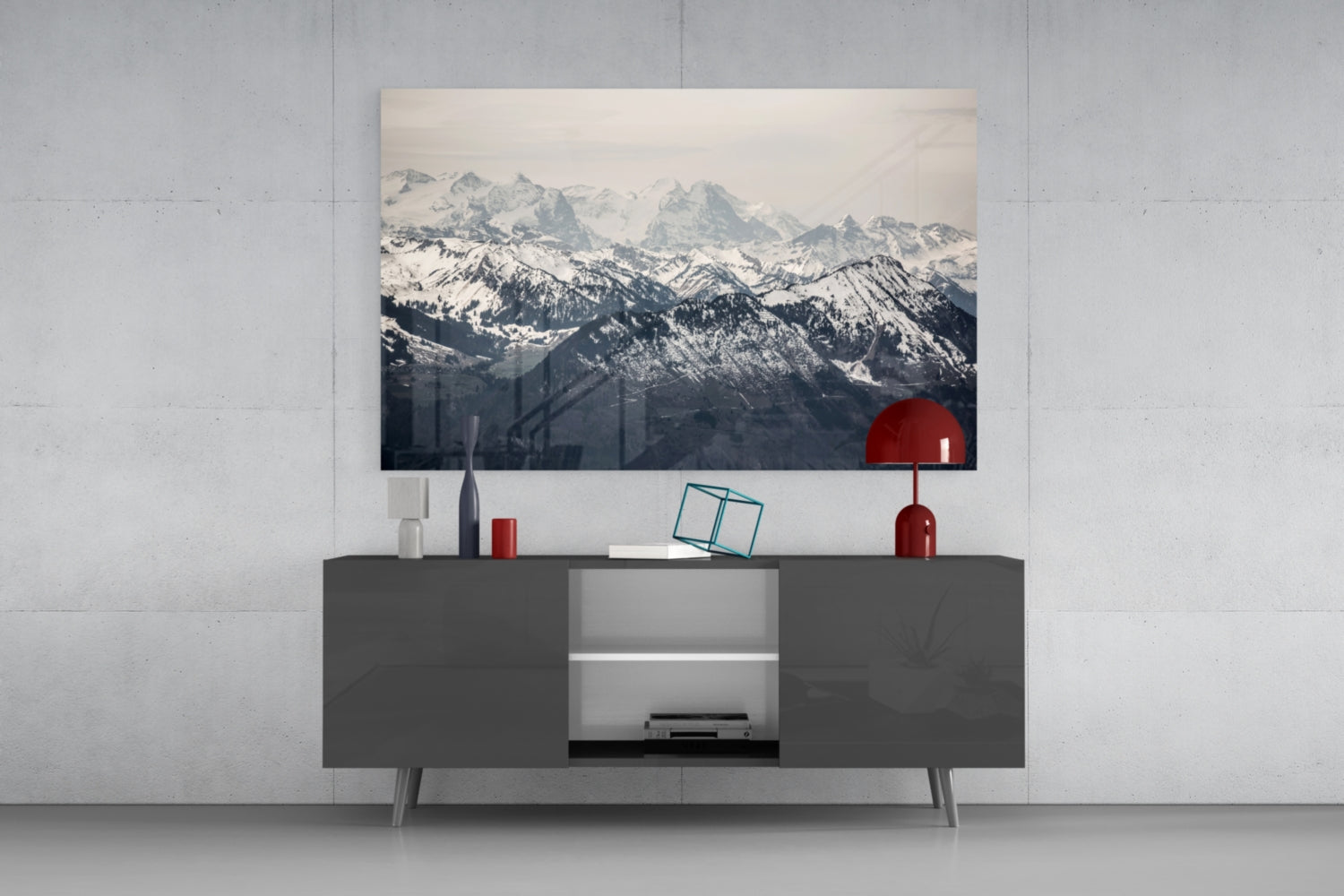 Snowy mountain landscape glass wall art displayed above dark gray media console with red decorative accessories and lamp.