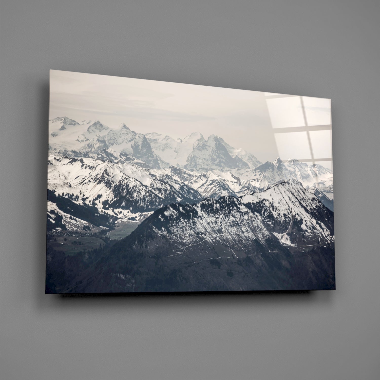Glass wall art mounted on gray wall displaying layered snow-capped mountain ranges with forested slopes and window reflections.