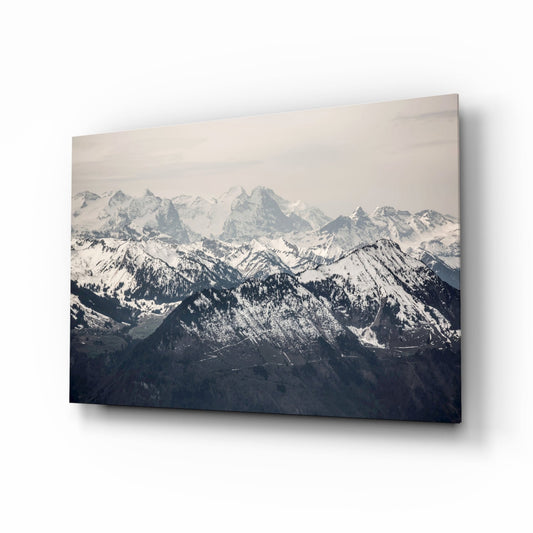 Glass wall art mounted on white background displaying snow-covered mountain range with forested foothills in muted tones.