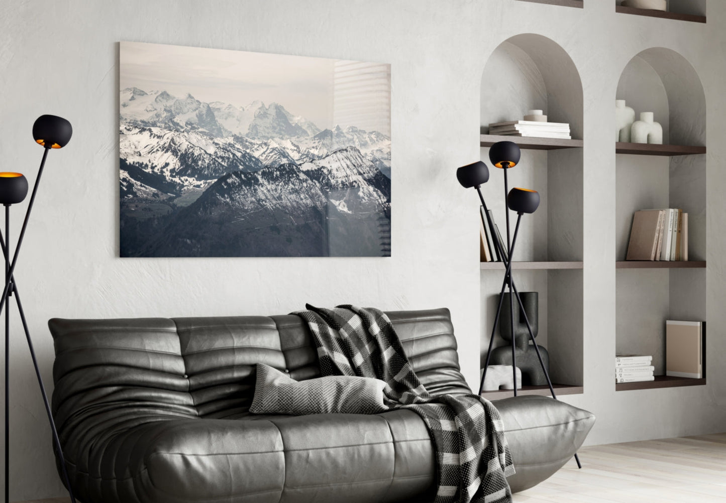 Glass wall art depicting snow-covered mountain peaks displayed above a black leather sofa in a modern living room setting.