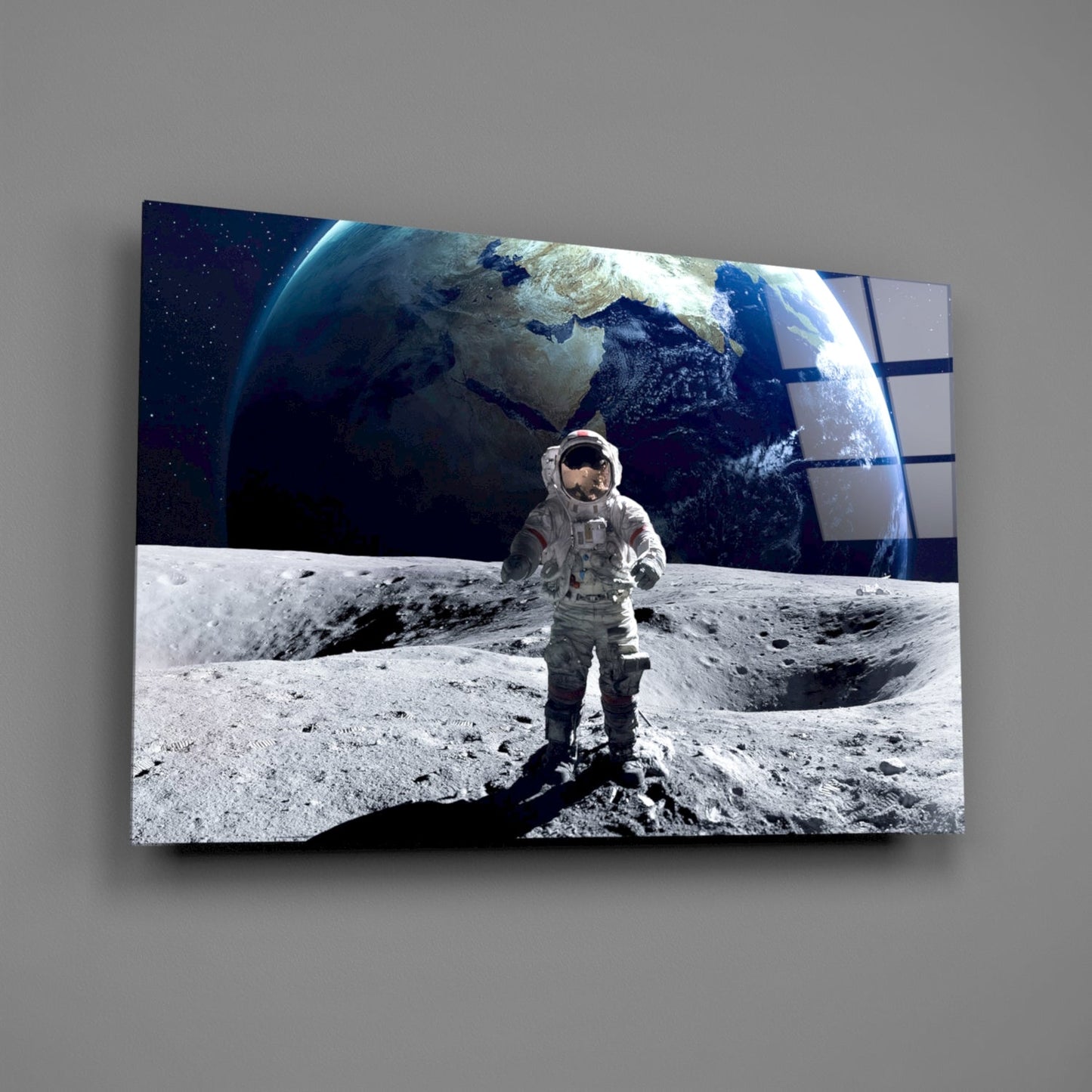 Astronaut standing on lunar surface with Earth rising above horizon, displayed as angled glass wall art panel against gray background.