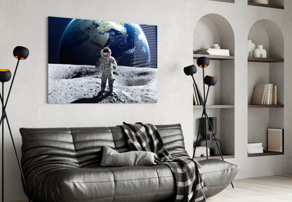 Astronaut on lunar surface with Earth in background displayed as framed glass art above black leather sofa in modern living room.