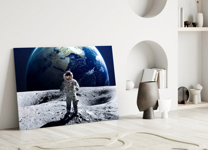Glass wall art featuring an astronaut on the lunar surface with Earth in the background, displayed leaning against a white wall in a minimalist interior.