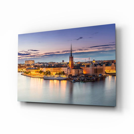 Glass wall art print displaying Stockholm's illuminated waterfront at dusk, featuring a church spire and buildings reflected in calm water.