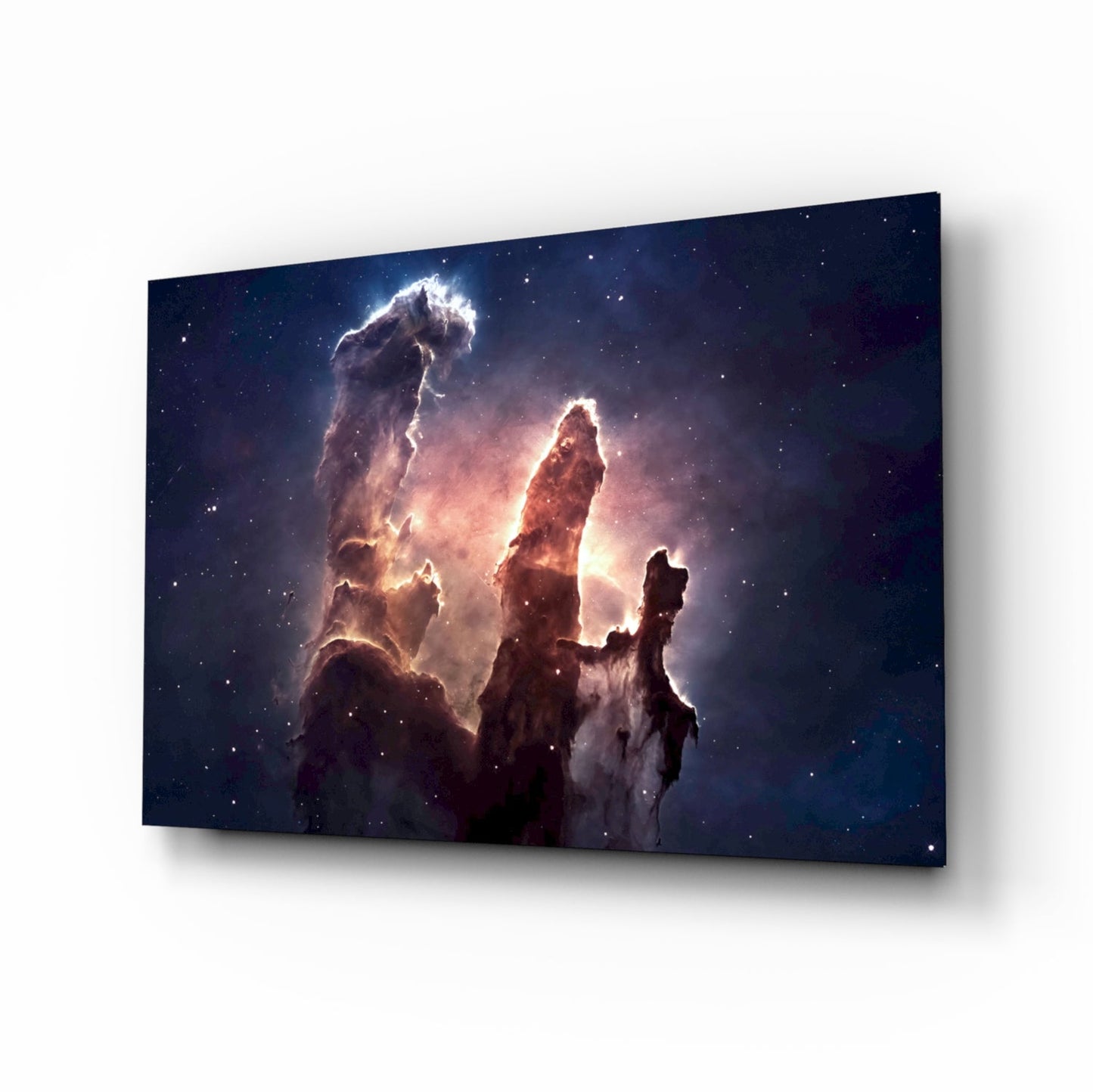 Glass wall art panel displaying the Pillars of Creation nebula with illuminated dust clouds in deep blue and amber tones against a starry background, photographed at an angle showing its three-dimensional depth.