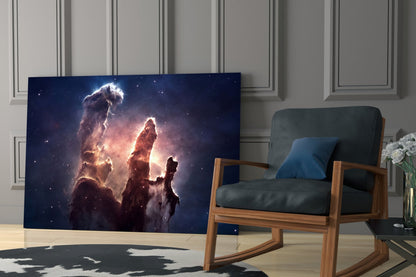 Space Clouds Glass Wall Art displaying a nebula with glowing pillars of dust and stars, displayed in a modern living room next to a wooden chair.
