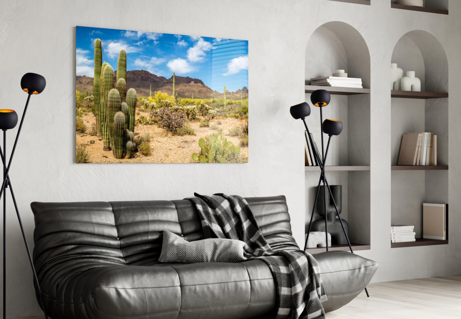 Glass wall art displaying a desert landscape with tall saguaro cacti, mountains, and blue sky, hung above a black leather sofa.