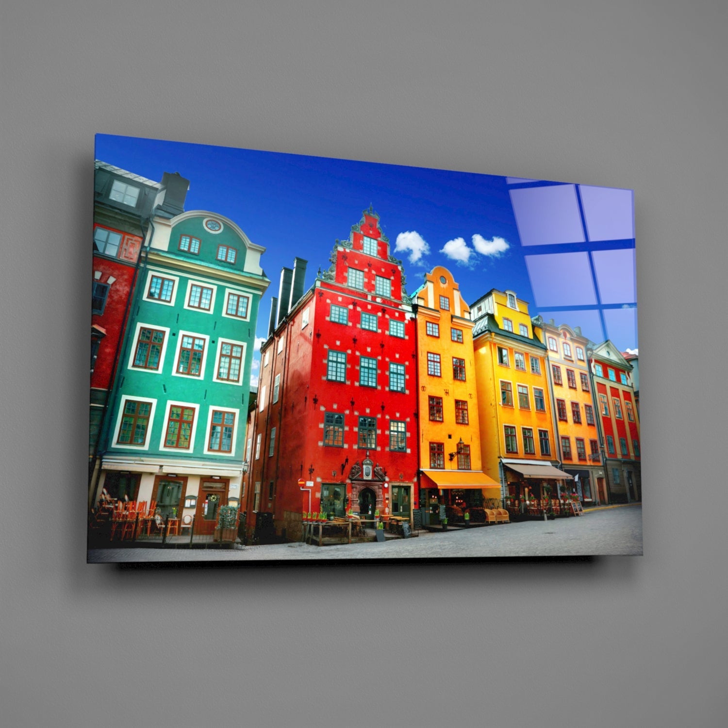 Glass wall art mounted on gray wall displaying colorful historic Stockholm buildings in red, green, and yellow under blue sky.