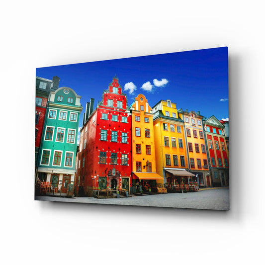 Colorful historic buildings in red, yellow, green, and orange with distinctive architecture displayed on a glass wall art panel against white background.