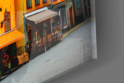 Angled view of glass wall art displaying a vibrant orange building facade with red flowers and wooden chairs.