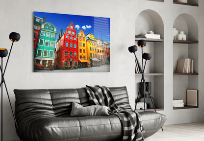 Glass wall art displaying colorful historic Stockholm buildings in red, yellow, and green hung above a black leather sofa in a modern living room.