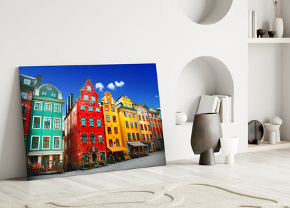 Glass wall art leaning against white wall displaying colorful historic Stockholm buildings in red, yellow, and green under blue sky.