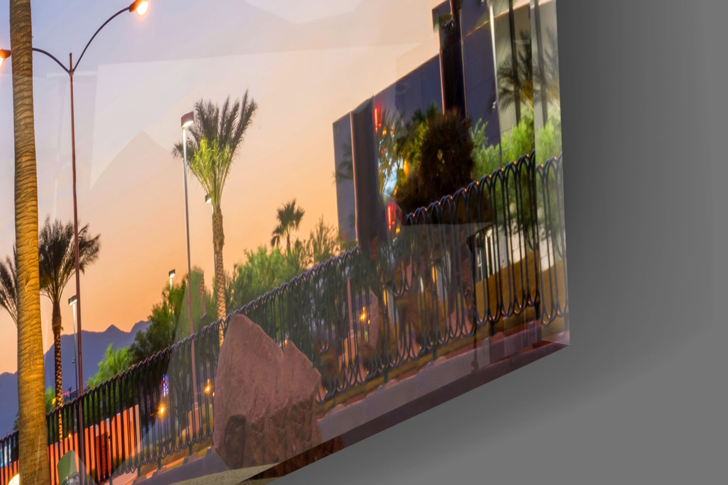 Angled view of illuminated glass wall art depicting palm trees and mountains at dusk with decorative wrought iron fence detail.