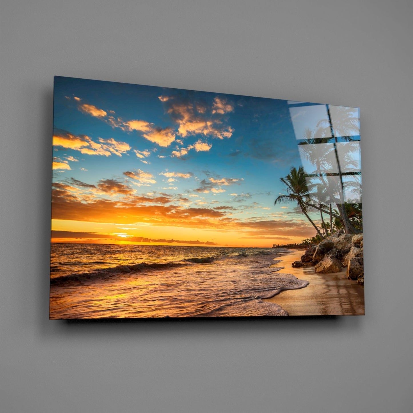 Beach sunset glass wall art mounted on gray wall, showing tropical shore with golden sky, ocean waves, and palm trees reflected in glass panel.