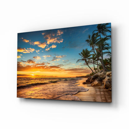Tropical beach sunset scene with palm trees and golden sky displayed on angled glass wall art panel.