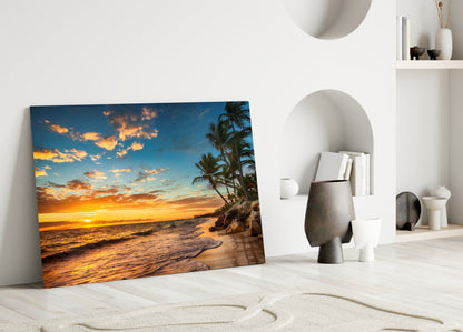 Beach Cruise Glass Wall Art leaning against white wall, displaying tropical sunset with golden sky, palm trees, and ocean waves.