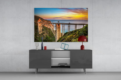 Bixby Creek Bridge glass wall art displayed above a dark gray media console, showing a sunset coastal landscape with a concrete bridge spanning cliffs.