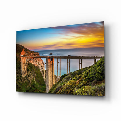 Bixby Creek Bridge glass wall art displayed at an angle, showing a dramatic coastal sunset with the iconic concrete bridge spanning a verdant canyon above turquoise ocean waters.