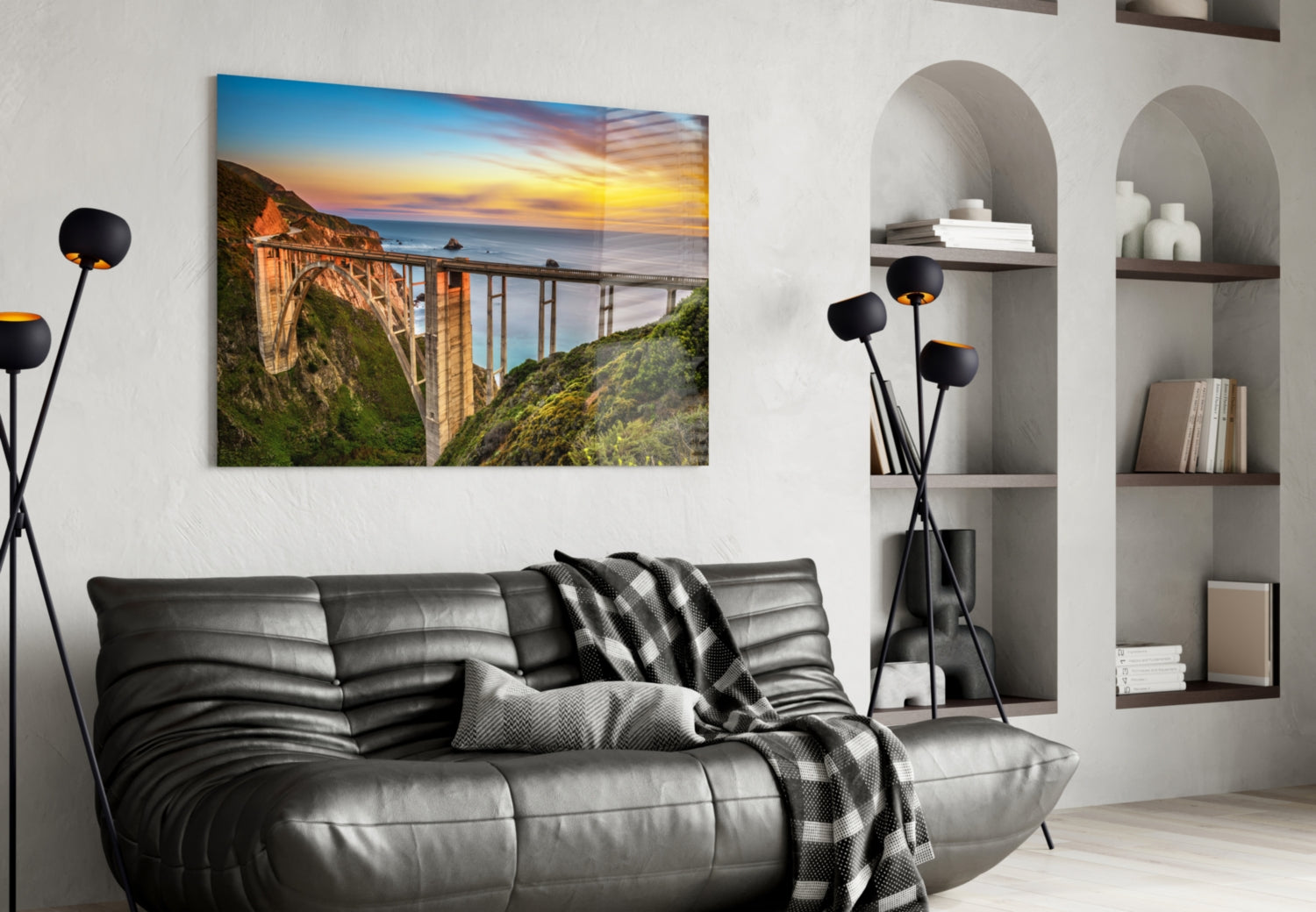 Bixby Creek Bridge glass wall art displaying a sunset coastal landscape hanging above a black leather sofa in a modern living room.