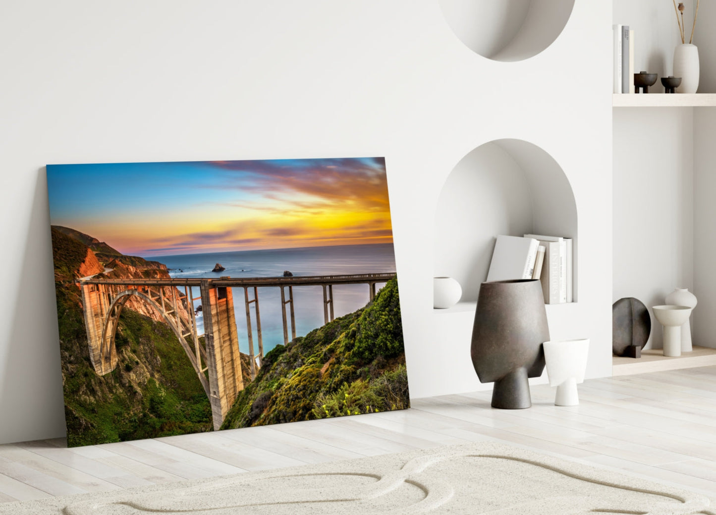 Bixby Creek Bridge glass wall art leaning against white wall in minimalist room with sand and decorative objects.