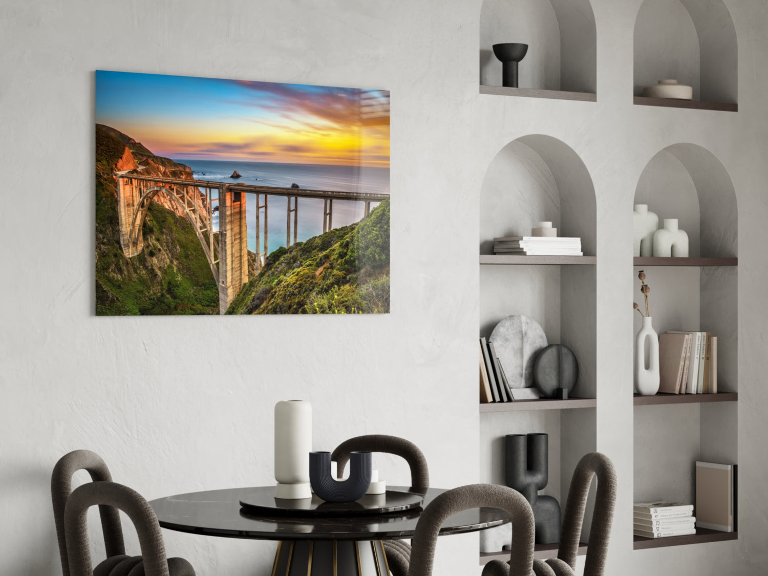 Glass wall art print of Bixby Creek Bridge at sunset displayed above a round black dining table in a modern interior.