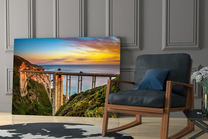 Glass wall art displaying Bixby Creek Bridge at sunset, positioned in a modern living room beside a wooden chair.
