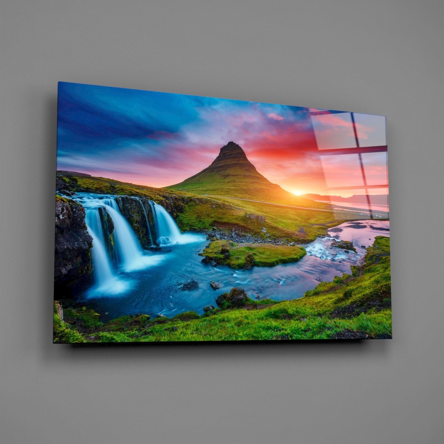Glass wall art mounted on gray wall displaying Icelandic waterfall landscape with mountain peak, blue water, green moss, and sunset sky.