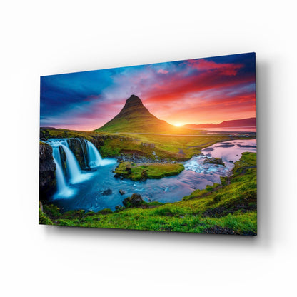 Glass wall art panel displaying Kirkjufellsfoss waterfall with green moss-covered landscape, blue water, and sunset sky in perspective view.