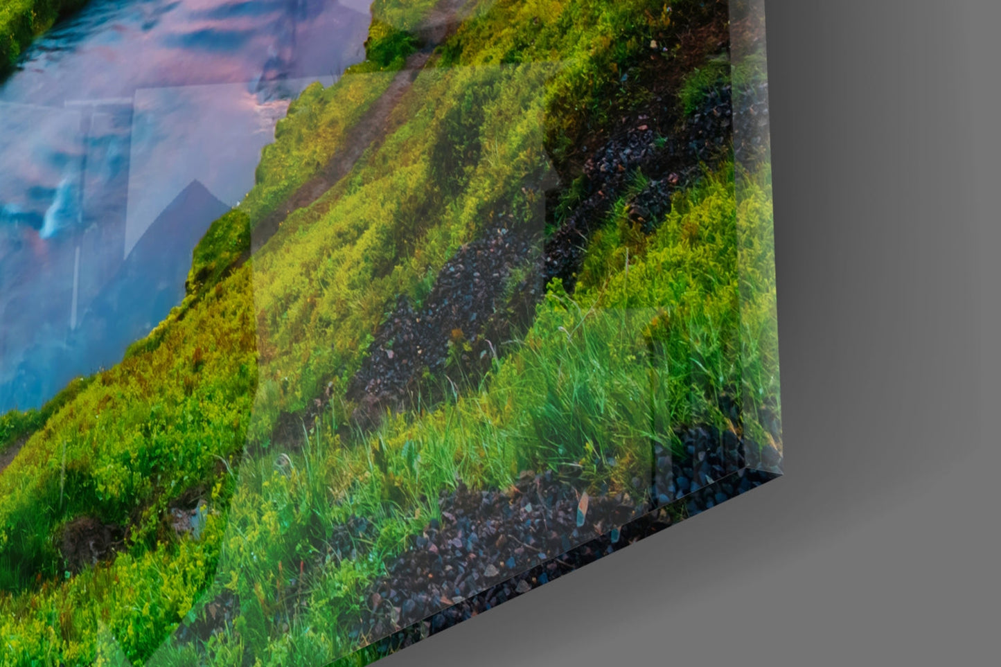 Close-up angled view of glass wall art showing lush green moss-covered hillside sloping toward blue water with mountain silhouette.