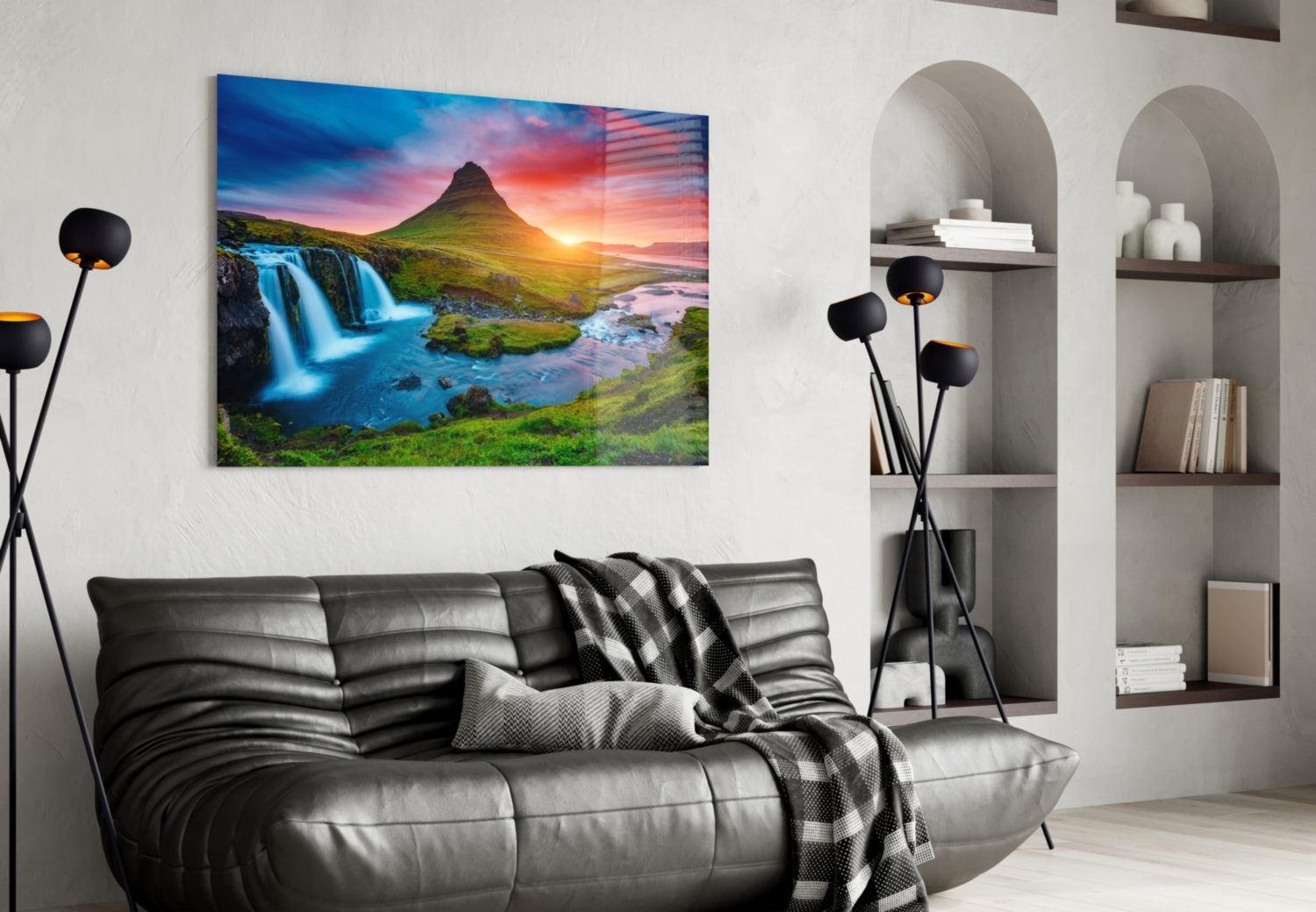 Glass wall art of Kirkjufellsfoss waterfall and mountain at sunset displayed above black leather sofa in modern living room.