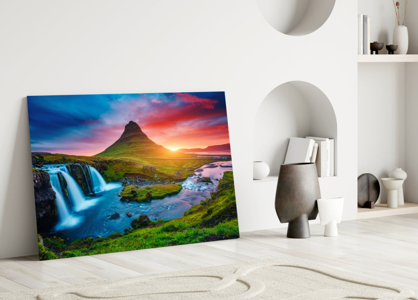 Glass wall art leaning against white wall displays Kirkjufellsfoss waterfall at sunset with blue water, green moss, and mountain peak.
