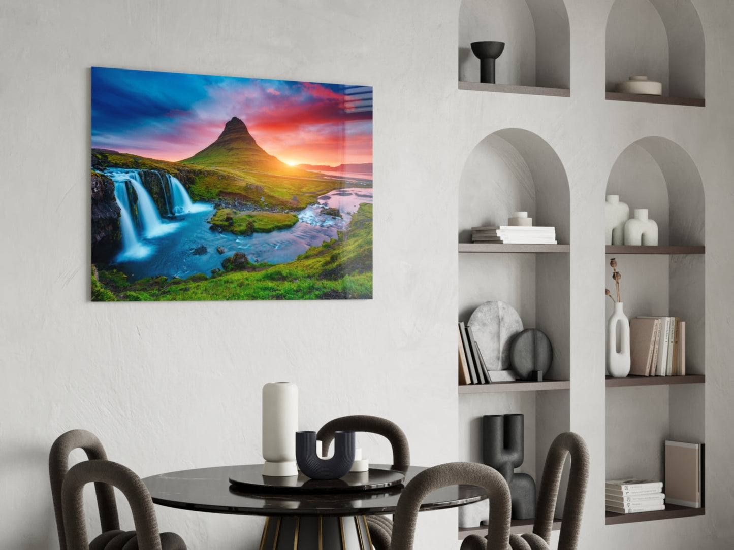 Glass wall art of Kirkjufellsfoss waterfall at sunset displayed above a round black dining table in a modern interior.