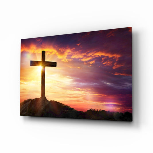 Silhouetted wooden cross on hillside against dramatic golden-orange sunset sky with purple clouds, displayed as framed glass wall art.