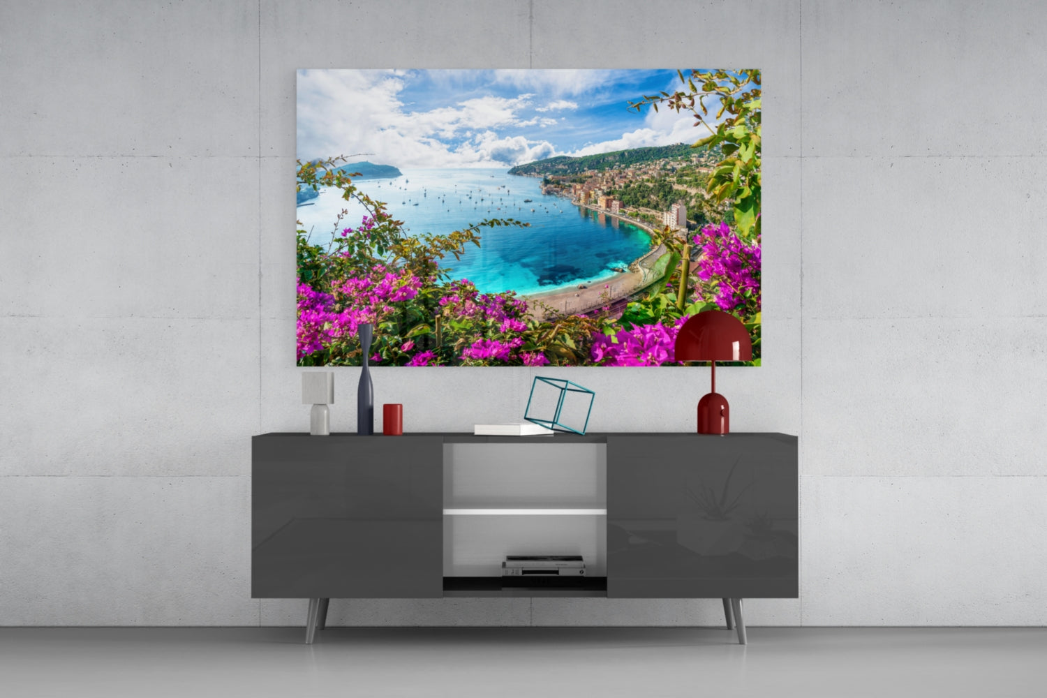 Glass wall art depicting Mediterranean coastal landscape with magenta flowers, turquoise sea, and sailboats, mounted above dark TV console.