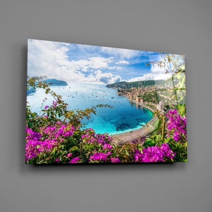 Glass wall art displaying a coastal Mediterranean landscape with vibrant magenta flowers, turquoise bay, sailboats, and hillside town.
