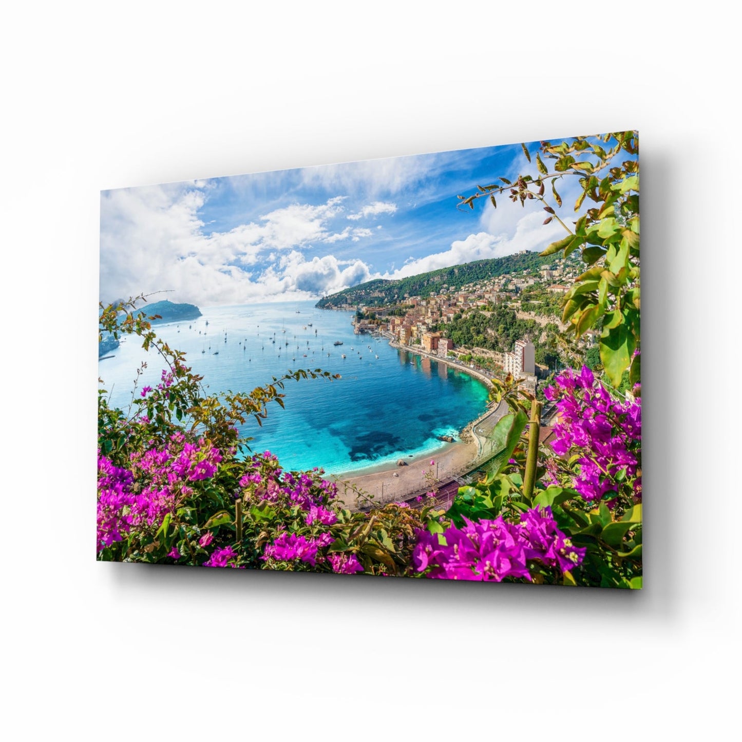 Glass wall art panel displaying a coastal Mediterranean landscape with vibrant magenta bougainvillea flowers framing a turquoise bay and hillside town.