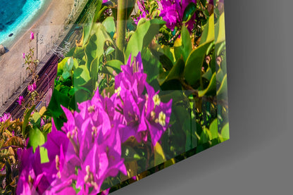 Glass wall art panel displaying vibrant magenta bougainvillea flowers with green foliage overlooking a turquoise Mediterranean beach.