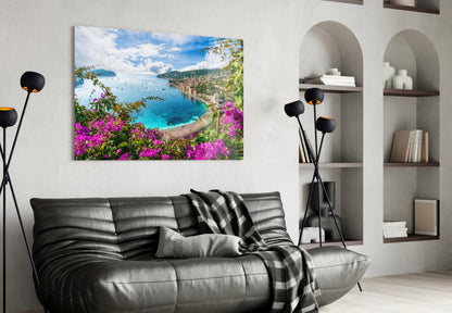 Glass wall art depicting a coastal Mediterranean bay with magenta bougainvillea flowers, turquoise water, and green hillside, displayed above a black leather sofa.