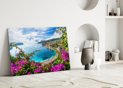 Glass wall art leaning against white wall displays vibrant coastal landscape with magenta flowers, turquoise sea, and blue sky.