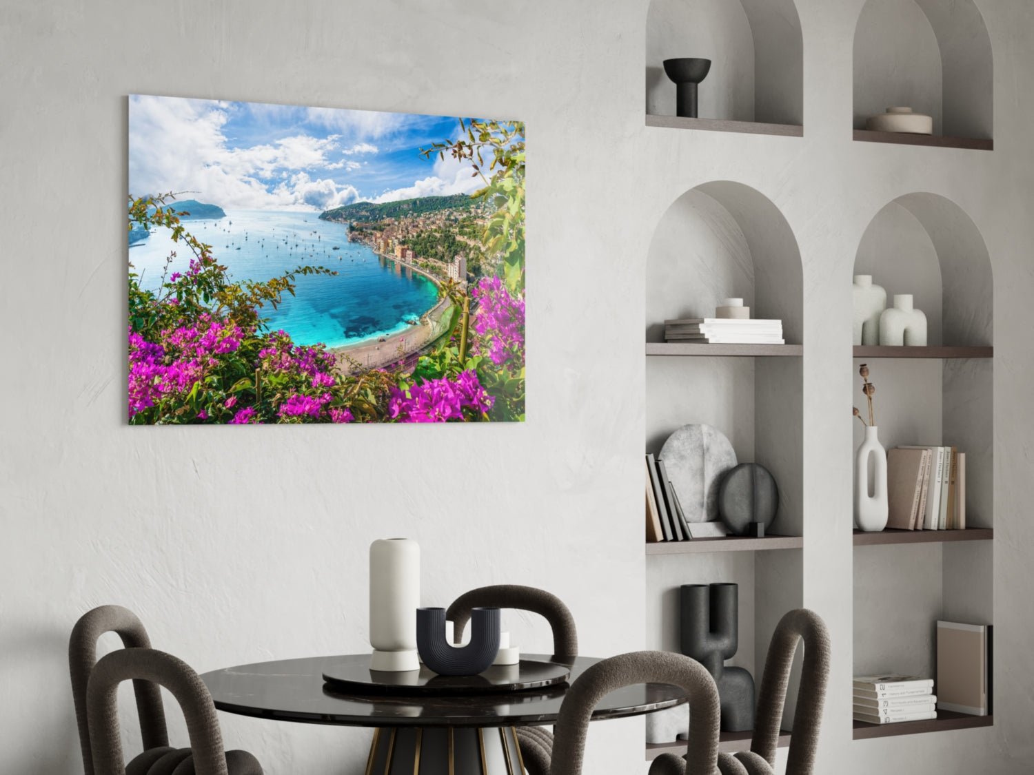 Glass wall art depicting a Mediterranean coastal bay with turquoise water, pink bougainvillea flowers, and green hillsides, displayed above a round black dining table.