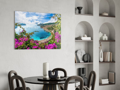 Glass wall art depicting a Mediterranean coastal bay with turquoise water, pink bougainvillea flowers, and green hillsides, displayed above a round black dining table.